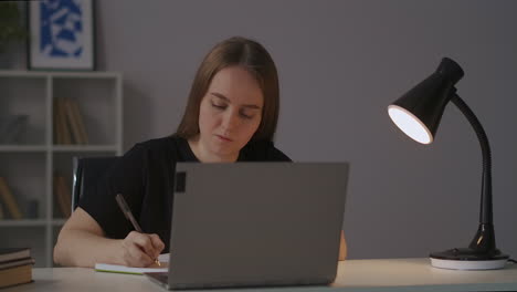 e-learning-at-home-female-student-is-viewing-lecture-online-and-writing-notes-in-notebook-portrait-of-woman-at-table-at-evening-table-lamp-is-lighting